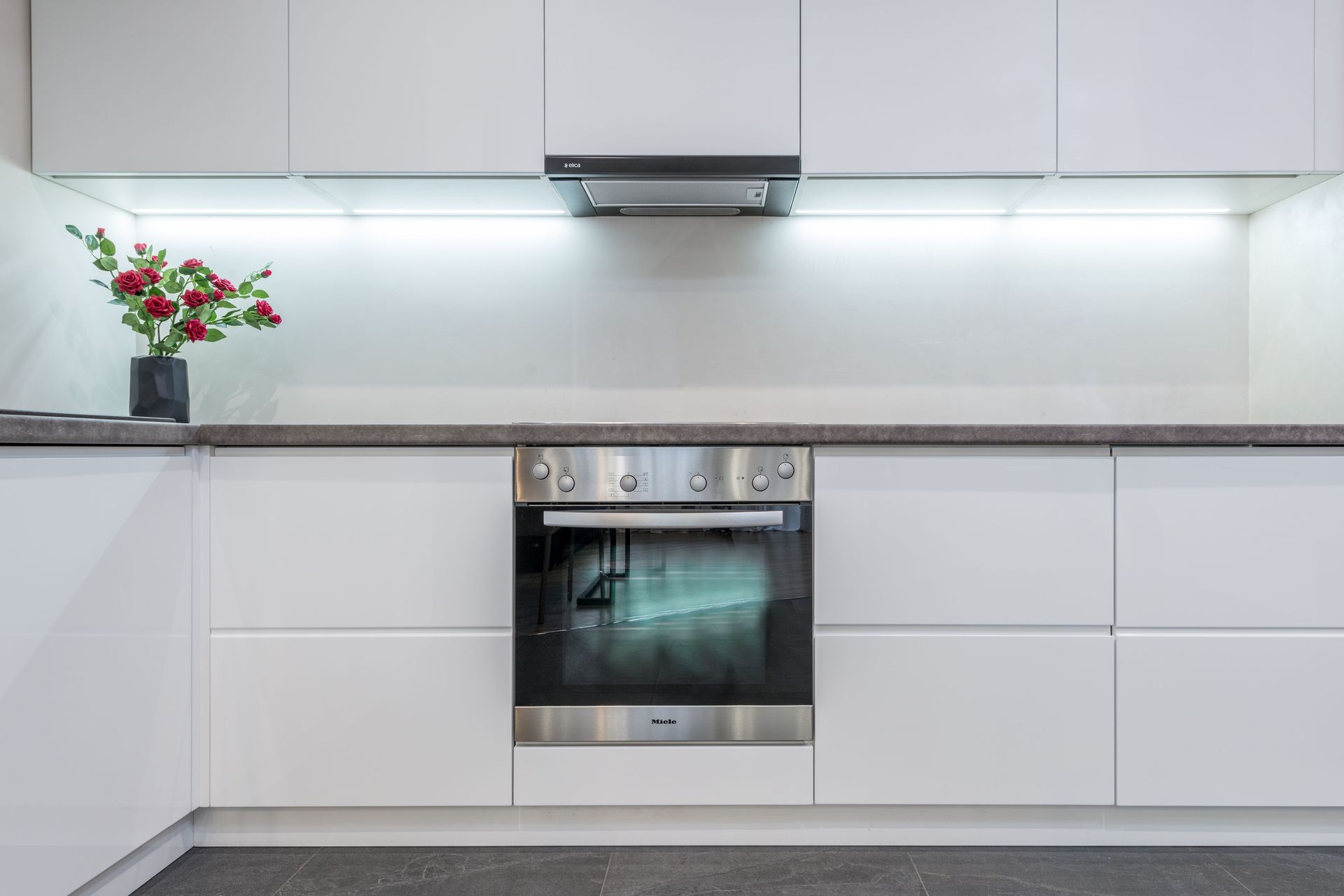 Modern chrome built in oven and hood in spacious illuminated kitchen with minimalist white cabinets decorate with fresh flowers in vase