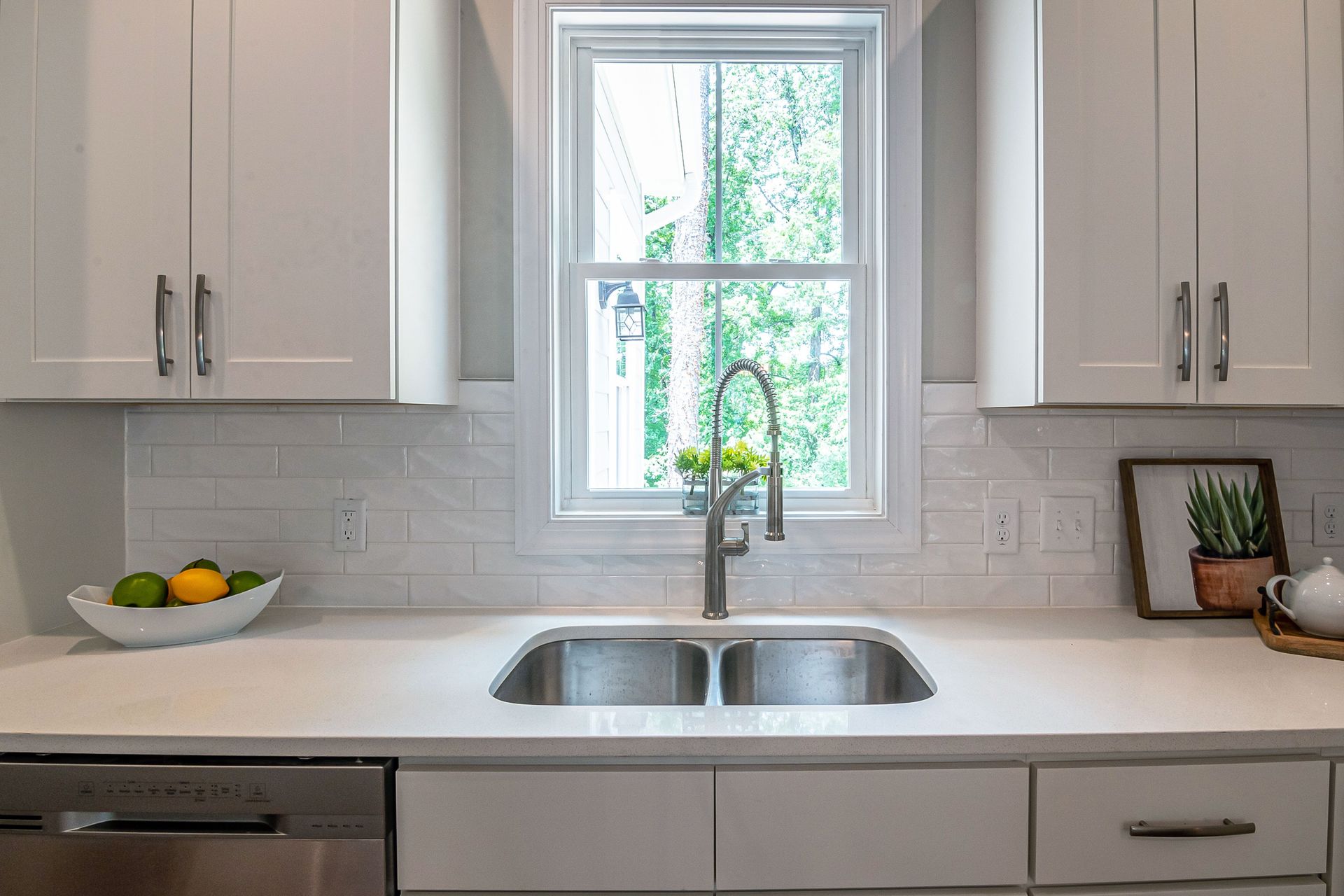 A contemporary kitchen with white cabinets, stainless steel sink, and lush outdoor view.